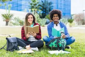 Diverse young couple holding books sitting lawn sensory friendly university campus.