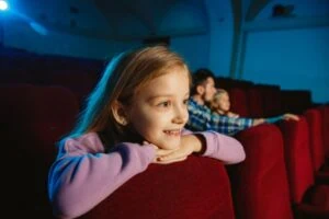 Young girl sitting in movie theatre leaning on chair.
