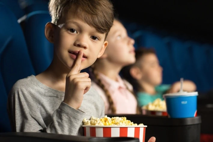 Young boy sitting in movie theatre with with his finger over his mouth.