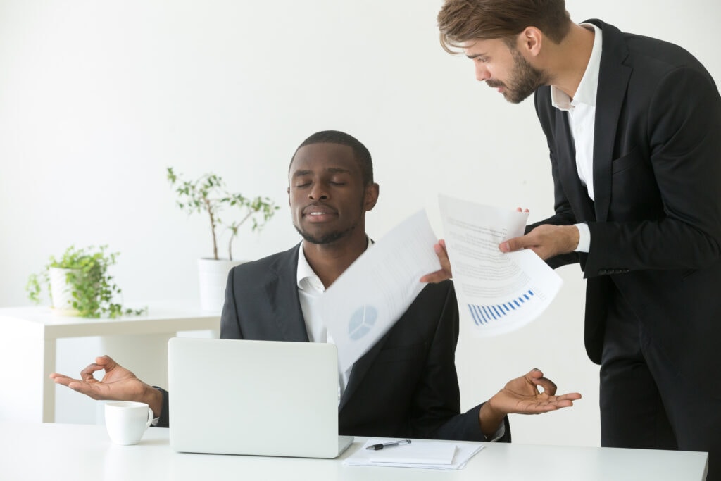 An african american person meditating at their desk at work, practicing mental health for startups, as someone brings the person papers to read.