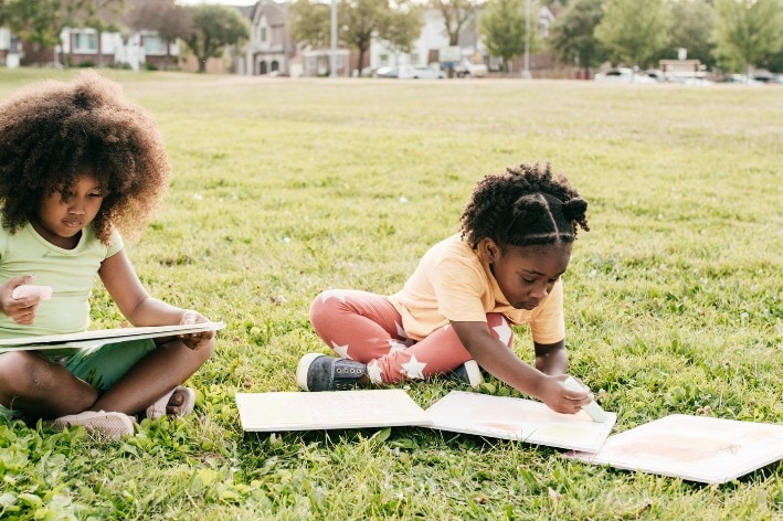 Two girls sitting on grass drawing on paper.