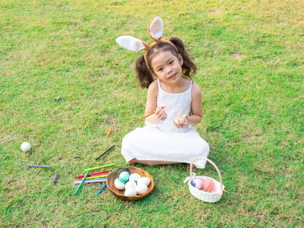 Little girl sitting on grass wearing bunny ears and decoaring an Easter Egg