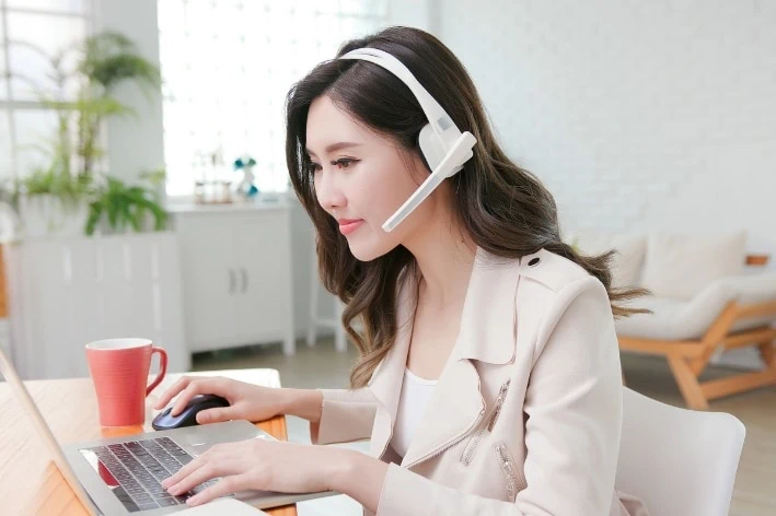 Young asian person sitting at desk in front of computer wearing a headset. 