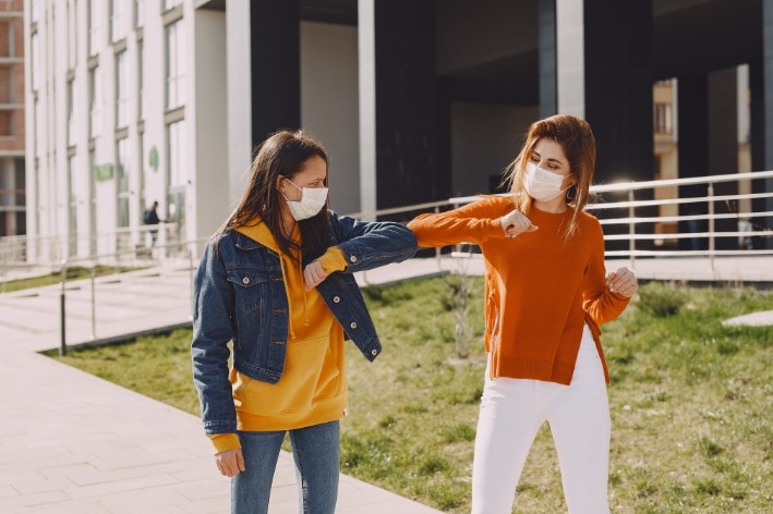 Two women walking wearing masks bumping elbows during COVID.