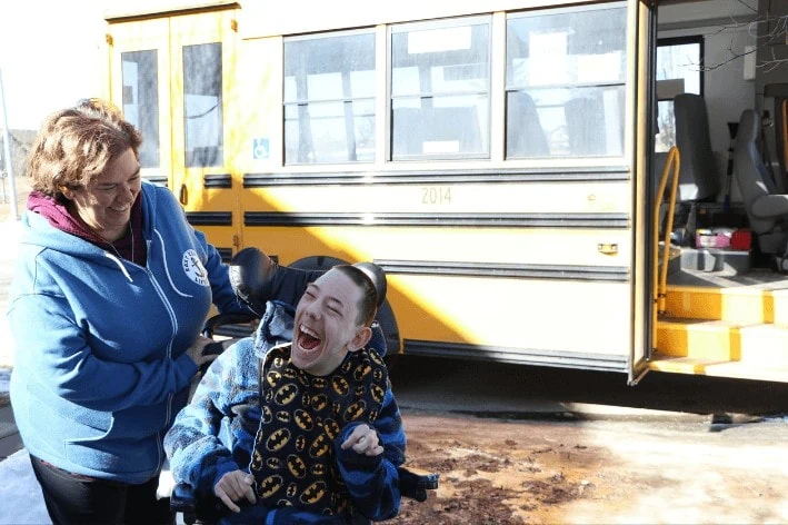 Male student in front of school bus in wheel chair being assisted by employee in front of sensory-friendly school bus.
