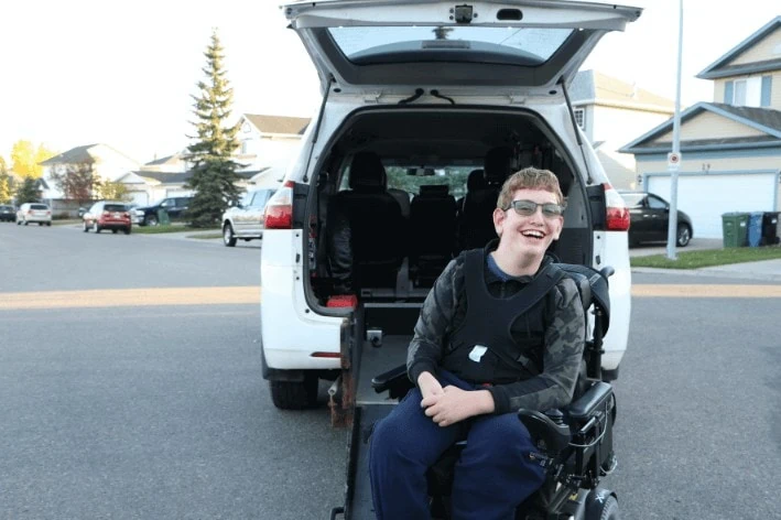 Student in wheelchair sitting in front of van with trunk open smiling at camera.