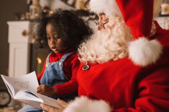 Young girl sitting beside Santa reading him a book.