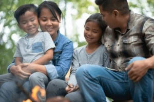 A young family of four sitting around a camp fire smiling.
