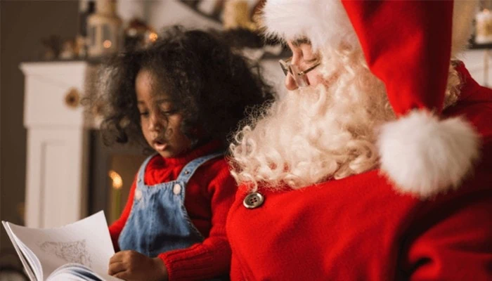 Young girl in overalls sits in Santa's lap. They are looking at a book together.