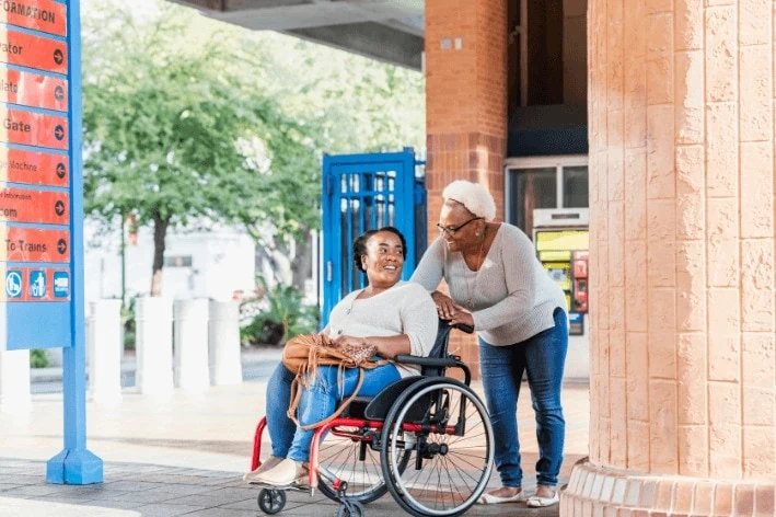 Person pushing another one in a wheelchair at a train station smiling at each other.
