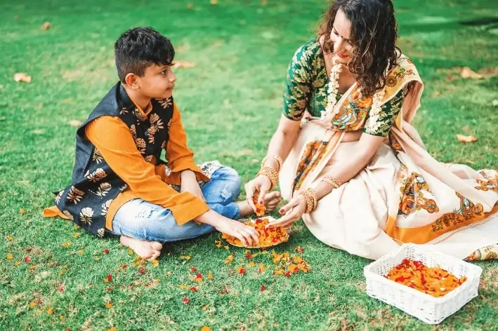 Adult and a child wearing cultural outfits sitting on the grass, playing with flowers.