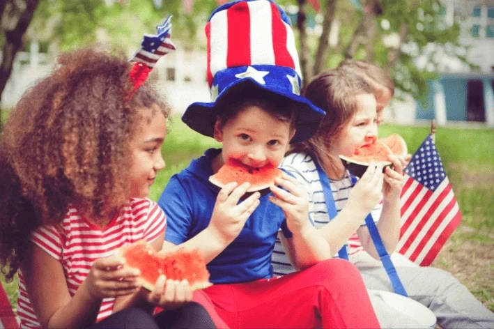 Children dressed up for Independence Day sitting on lawn eating watermelon.