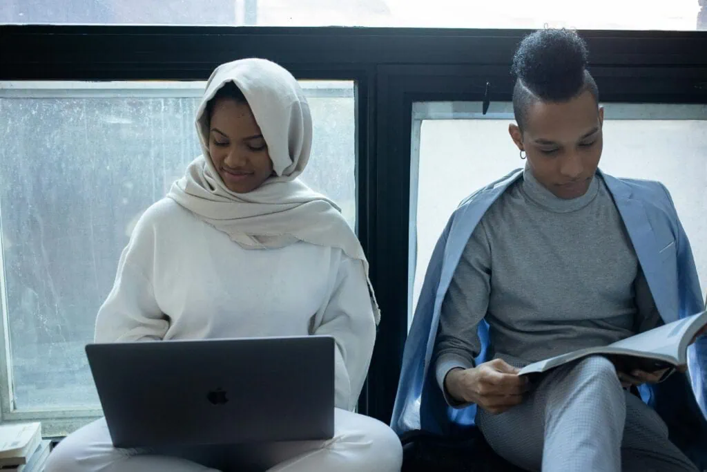 A woman wearing a head scarf typing on her laptop, a man making notes on a notepad