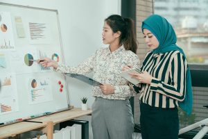 two women standing near a white board with business data on it one woman is asian wearing a dress shirt and trousers the other woman is wearing a teal hijab and a striped dress shirt with black trousers